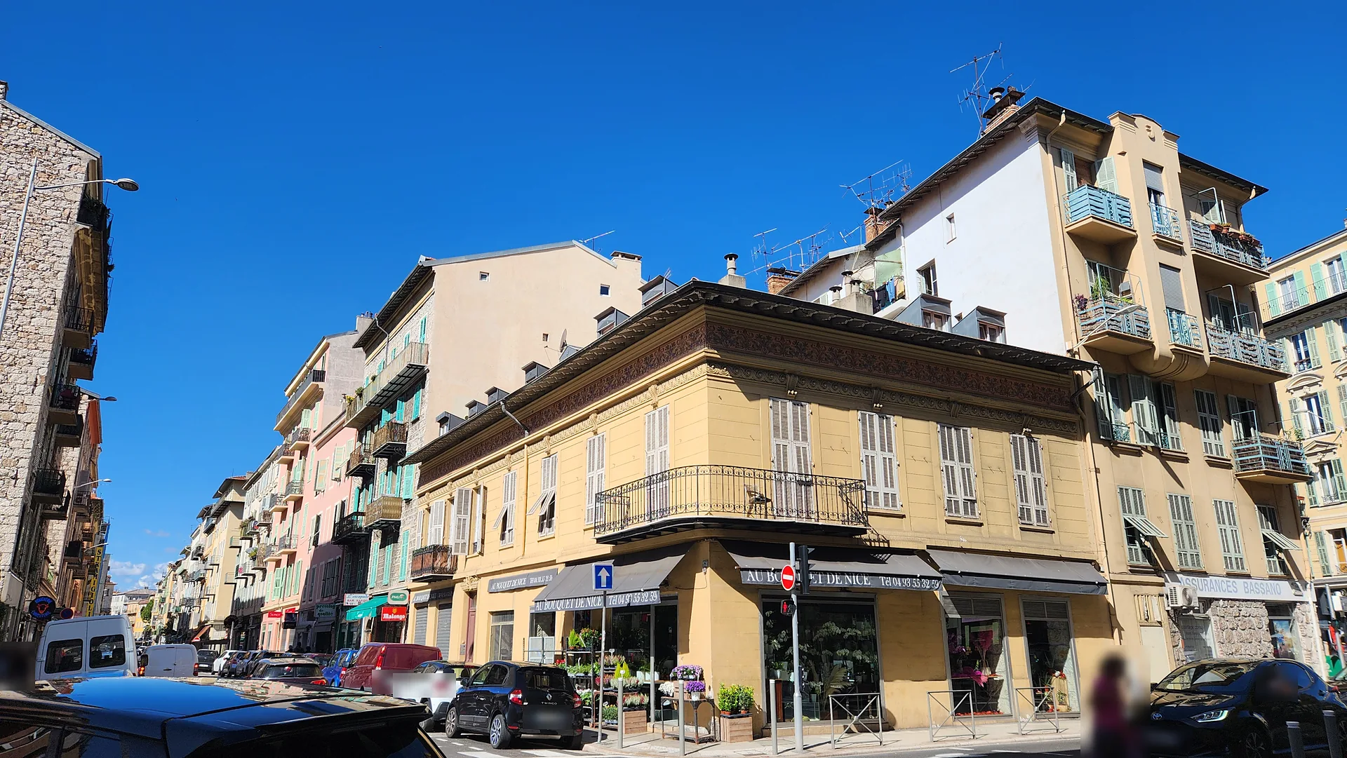 Petit immeuble ancien jaune à balcon d'angle en fer forgé, avec un fleuriste au rez-de-chaussée, devant des immeubles plus hauts